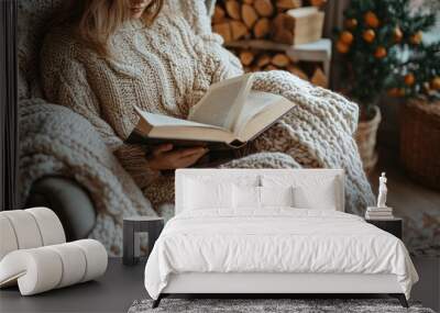 A woman in a cozy knitted sweater sits in an armchair by a fireplace and reads a book. Wall mural