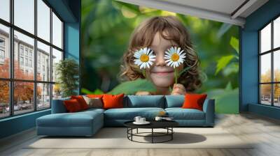 young girl, possibly in her early childhood years, playing in a lush green outdoor setting. She is holding two white daisies close to her eyes, playfully obscuring her vision Wall mural