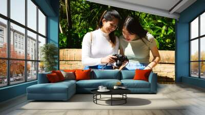 Two Asian women on holiday. They are sitting in a park looking at the pictures on the camera. On her legs she is holding a tourist map of the city. Two young women are happy looking at the pictures. Wall mural