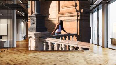 young and pretty brunette Moroccan woman dressed in white top and jeans is happy leaning on the railing in the square of Spain in Seville, she looks back and takes the photo. Wall mural