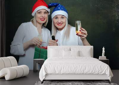 two joyful smiling girls in bright  santa hats, with a gift in hand and white outfits having fun drinking champagne covered in a craft bag, celebrate the new year. Wall mural