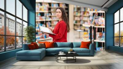 A young beautiful woman poses with a book in her hands, against the background of shelves with books. The concept of education and purchase of books Wall mural