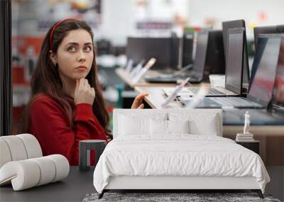 A young brunette woman in a red sweater thoughtfully selects a laptop in the store. Close up. The concept of buying digital goods Wall mural