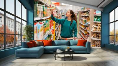 A young Caucasian beautiful woman with a grocery cart, takes a box of juice from the top shelf. Shelves with goods in a blur in the background. The concept of buying goods and shopping Wall mural