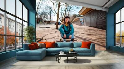 A young Caucasian woman is sitting cross-legged on a skateboard. In the background, a street with trees. Copy space. Concept of sports lifestyle and street culture Wall mural