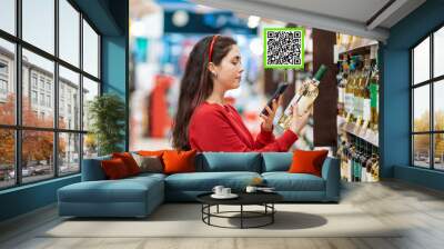 A young Caucasian woman scans the qr code on a bottle of wine using her smartphone. Qr code icon above the product. In the background is a supermarket. Concept of modern technologies and shopping Wall mural