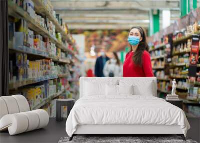 A young woman in a medical mask poses in the aisle of a supermarket. The concept of consumerism and the new reality Wall mural