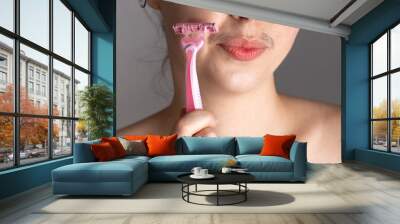 A young woman with a mustache winces, touching her face with a razor. The concept of getting rid of unwanted facial hair. Close up Wall mural