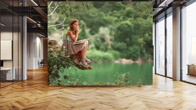 Pensive schoolgirl in a dress and a straw hat reading a book sitting on a old log. In the background are park and river. Copy space. The concept of children's education and school holidays Wall mural