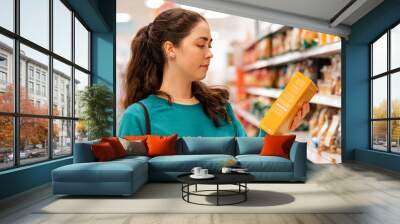 Portrait of a young Caucasian beautiful woman who reads the ingredients on the product packaging. Shelves with goods in a blur in the background. The concept of buying goods and shopping Wall mural