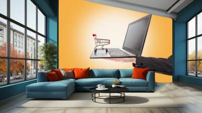 Shopping. A man's hand holds a laptop with a mini grocery cart standing on it. Orange background. The concept of online shopping Wall mural