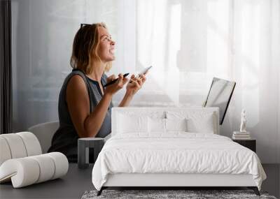 Stress at work. A young Caucasian woman screams in exasperation, holding a phone in her hands. There is a light window in the background. The concept of self-isolation, quarantine and remote work Wall mural