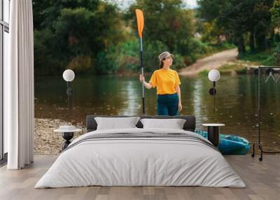 World Tourism Day. A young happy woman stands with an oar in her hand near the kayak. The concept of kayaking and outdoor activities Wall mural