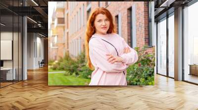 Сute happy red-haired woman stands near the apartment building where she lives and laughs at the camera. Space for text. Wall mural