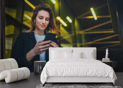 Successful female banker using smartphone outdoors while standing near his office background yellow neon lights, young woman professional manager working on mobile device near skyscraper at night Wall mural