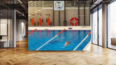 Outdoor swimming pool with clock on the wall to record the speed and time of the swim, near cardio equipment exercise bike and a life buoy. Wall mural