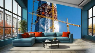 Scale active skyscraper building, glare from the sun on the windows, cranes against the blue sky. Wall mural