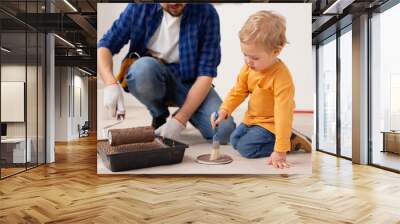 A close-up of a 2-year-old blond boy wearing an orange sweater and jeans paints the floor with a paintbrush. The child is very focused on what he is doing. His father is by his side. Wall mural