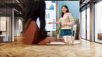 A focused young woman engaged in her work stands at a flipchart with charts and graphs, presenting market research results to company associates. The woman answers questions from the audience Wall mural