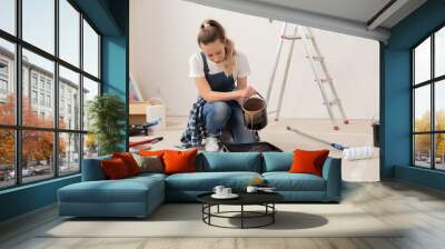 A teenage girl, dressed in a white T-shirt, denim overalls and a checkered shirt, squatted in the middle of the room. She pours brown paint into a plastic construction bowl. Wall mural