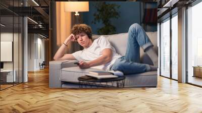 A young boy with curly hair lies on the couch at home watching TV with wireless headphones. Teenager listens to music,podcast,news while relaxing in the evening Wall mural