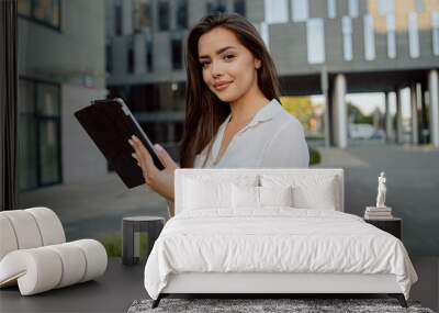 Attractive pretty girl in white shirt stands in front of glass modern business building of insurance corporation company, woman holds tablet in hand browses web writes email reads news Wall mural