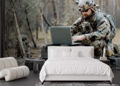 Bearded soldier in uniform sit on military transport crates, analyze data on a laptop and work out tactics at a temporary forest base. In the background, you can see a soldier protecting the base. Wall mural