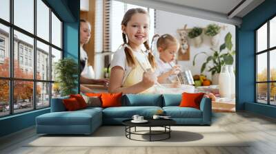 Close-up shot curious beautiful girl wearing apron kneading dough holding spoon in hand smiling at camera. In background little sister holding metal sieve looking in it and youn mother. Wall mural