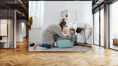 Mom plays with son who sits in laundry bowl, boy wants to spend time with woman and help her with household chores, putting clothes in washing machine Wall mural