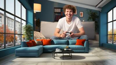 Portrait of smiling young boy, student with curly hair who is looking at the camera. The teenager is pressing buttons from a pad, playing on a console. Wall mural