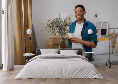 Smiling brunette with dark complexion is standing at window of new apartment, looking out at the view from it, drinking coffee in a mug, living room piled with cardboard boxes with things after move Wall mural