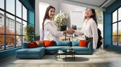 Two women elegantly dressed are standing in middle of corporate office, they are smiling at each other shaking hands in friendly manner, greeting, saying goodbye, getting acquainted, congratulating Wall mural