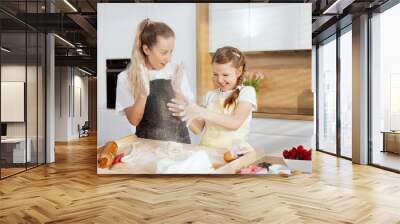 Young mom with schooler daughter clapping in hands with flour kneading dough preparing cookies biscuits pie using kitchen equipment rolling pin spoon knife. Wall mural