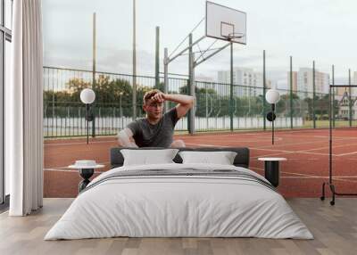 Young tired exhausted guy man in glasses wearing sportswear sitting at basketball court outside resting relaxing after training time doing exercises with company guys friends. Basketball game process. Wall mural