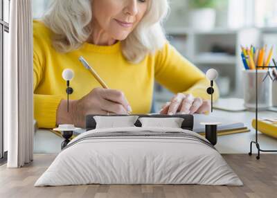 A close-up of a mature woman writing notes during an online course, accentuated by a modern home office setup. Wall mural