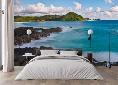 view of the vast blue sea with white waves hitting black coral rocks on the beach and next to it there are green hills filled with grass with blue skies and thick white clouds on Selong Belanak beach Wall mural