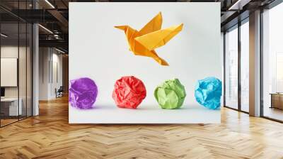 A photo of an origami bird flying over four differently colored crumpled paper balls on a white background, symbolizing creativity and imagination in business idea. Wall mural