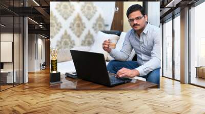 Young successful businessman working on laptop and filling tea cup with tea for refreshment after too much work. Wall mural