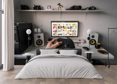 Cloud gaming: a gamepad in a male hand against the background of a gaming table and a monitor with a lot of games. Wall mural