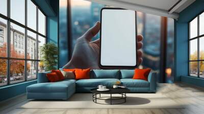 A person holds a blank smartphone against a blurry sunset view from a modern skyscraper window, displaying urban lifestyle. Wall mural