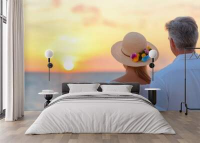 An elderly couple is standing on a beach, with the sun setting in the background. They are both wearing straw hats Wall mural