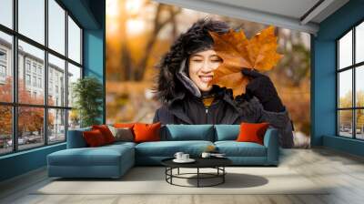 Asian young woman wearing a black sweater hoodie holding a falling brown maple leaf covering some part of her face in winter, headshot. Wall mural