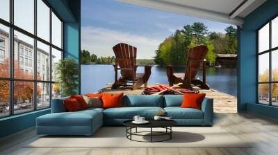 Two Adirondack chairs on a wooden dock facing the blue water of a lake in Muskoka, Ontario Canada. Canoe paddles and life jackets are on the dock. A cottage nestled between green trees is visible. Wall mural