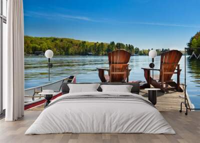 Two Adirondack chairs on a wooden dock on a lake in Muskoka, Ontario Canada. A red canoe is tied to the pier. Across the water cottages nestled between green trees are visible. Wall mural