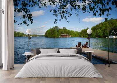 Two Adirondack chairs on a wooden dock overlooking a calm lake. A cottage nestled between green trees are visible across the water. Tree branches are framing the capture. Wall mural