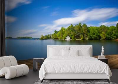 Wood dock in Muskoka facing a lake during a sunny summer day. Across the calm water there's a white cottage nested into the green trees.  Wall mural