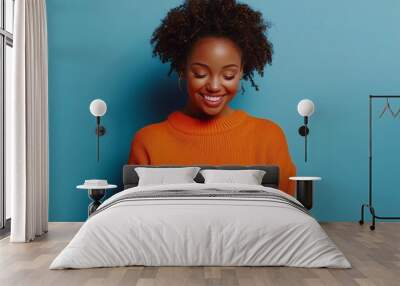 Photo of cheerful delighted african american woman types sms on modern cell phone device enjoys good internet connection dressed in orange jumper focused aside isolated on blue studio wall. Wall mural