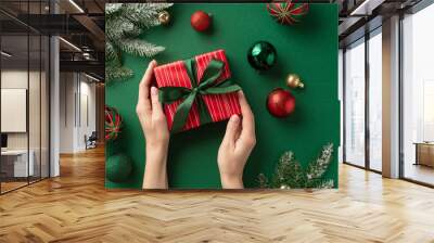 Christmas Eve concept. First person top view photo of woman's hands holding red gift box with ribbon bow over green gold red baubles balls and pine branches in frost on isolated green background Wall mural