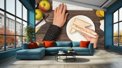 First person top view photo of female hands holding sandwich over plate and typing on keyboard mouse apples glass of juice flowerpot and stationery on isolated dark wooden table background Wall mural