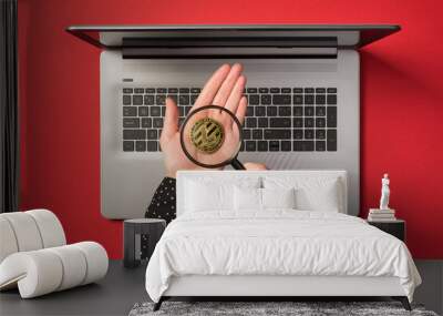 First person top view photo of hands holding gold coin with litecoin symbol and magnifying glass over open grey laptop on isolated red background Wall mural